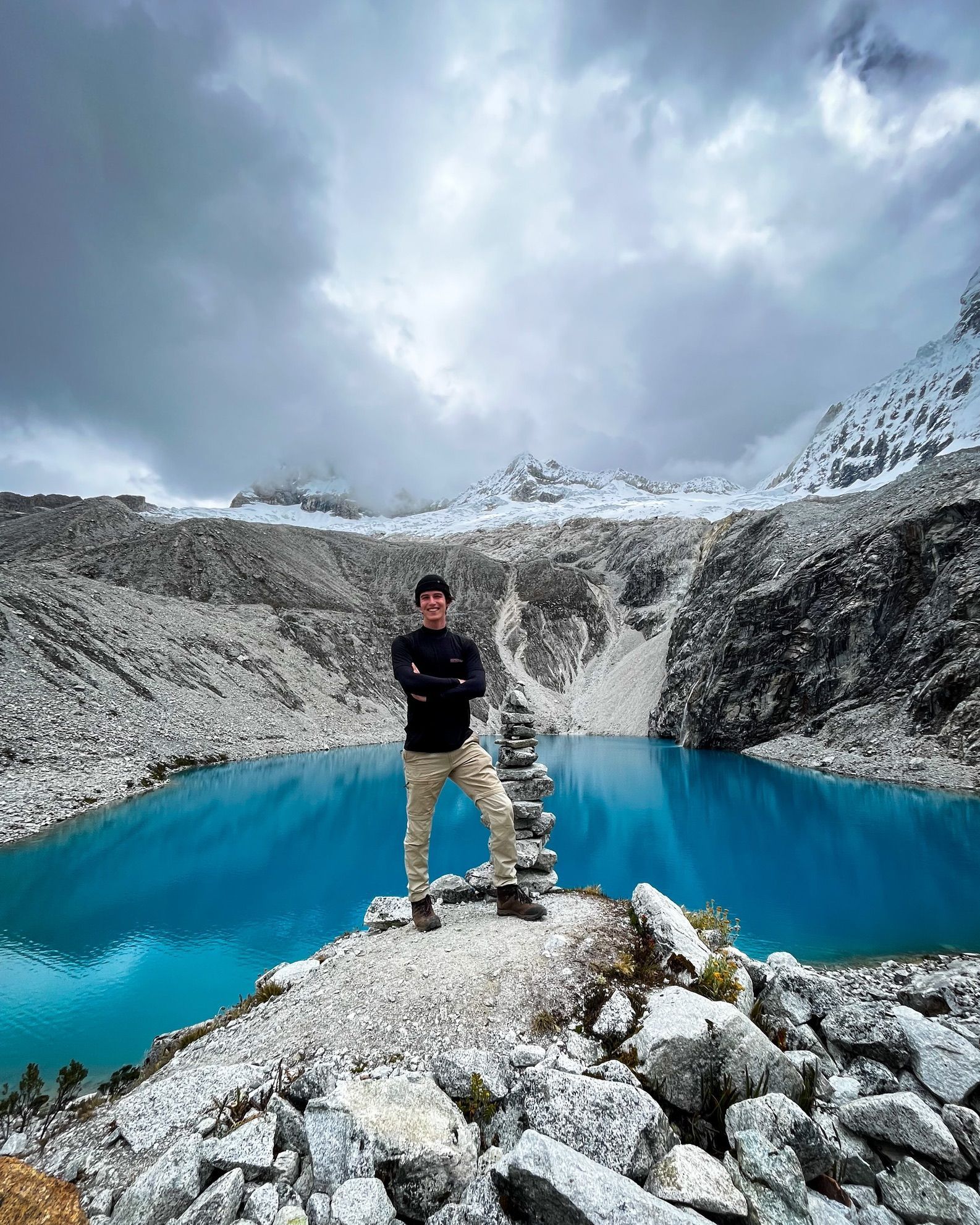 Tom in front of a turquoise glacial lake, snow peaks behind.