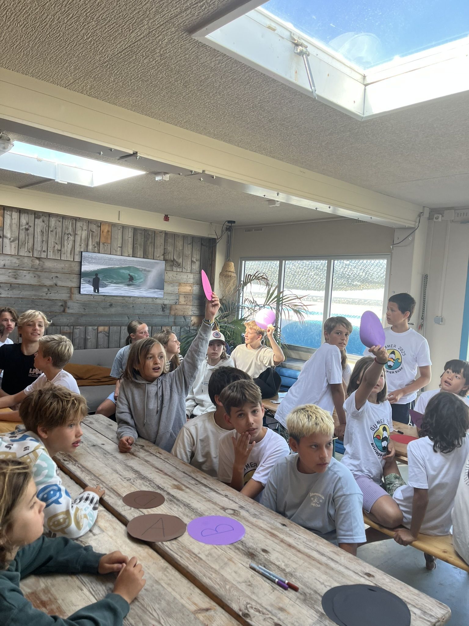 A surf camp room — kids around a long table, paddles raised, energy high.