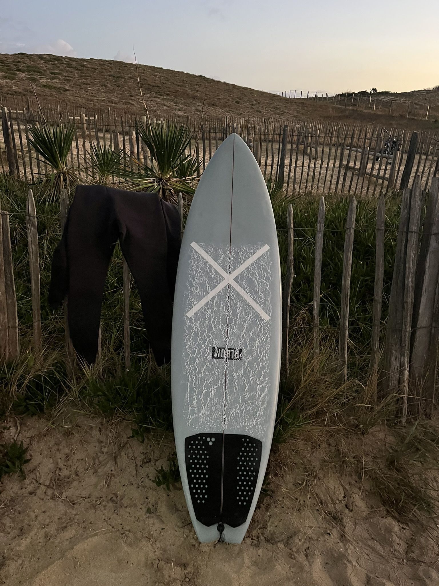 A surfboard standing against a wooden dune fence at sunset, wetsuit hanging.