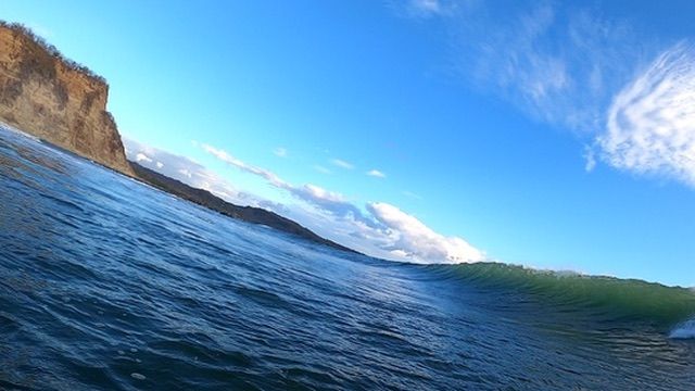 From the water — a wave rising in front of the camera, cliffs to the left.